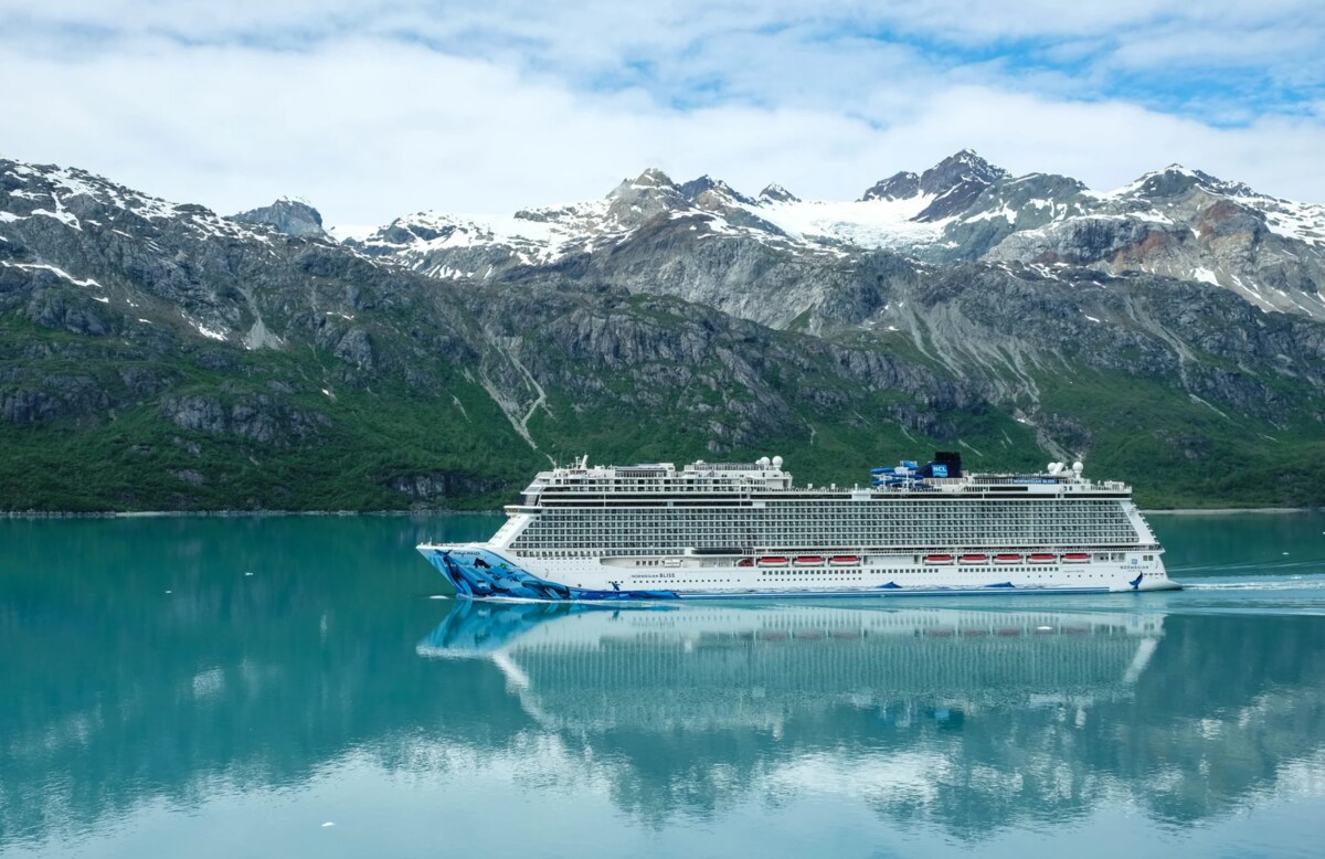 An ocean cruise ship sailing through turquoise water beneath snow-covered mountains in Alaska
