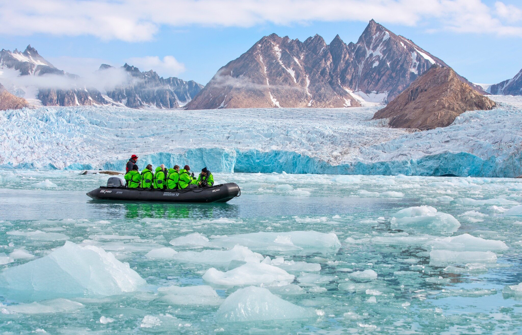 Expedition travelers crossing icy Arctic water by Zodiac beneath a glacier and jagged mountains