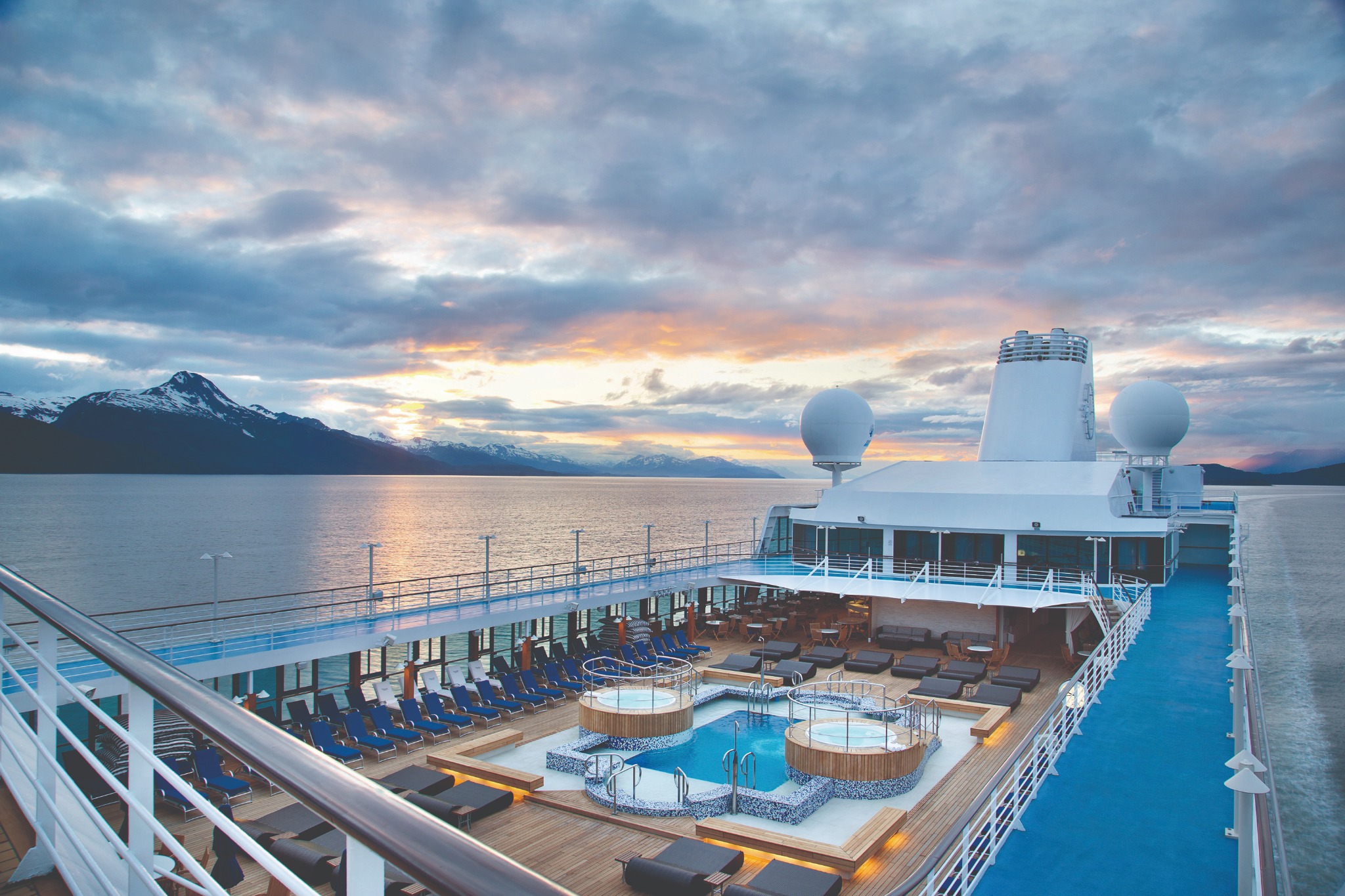 An ocean cruise ship’s pool deck beneath a colorful sunset and mountain-lined coast